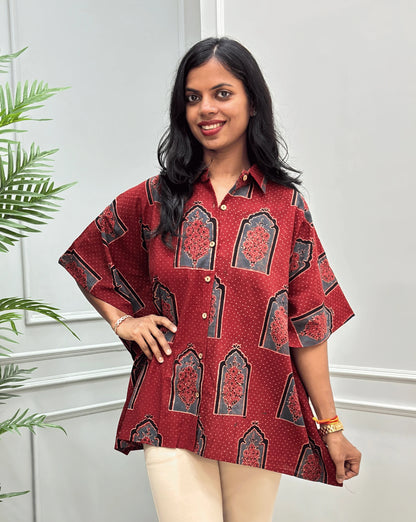 Front View of Red Women's Ajrakh Pure Cotton Short Kaftan Shirt with a Black, Grey & Red Floral Block Print, Wooden Button on front, paired with beige flared pants, standing on a round woven rug in front of a white paneled wall with a potted plant.