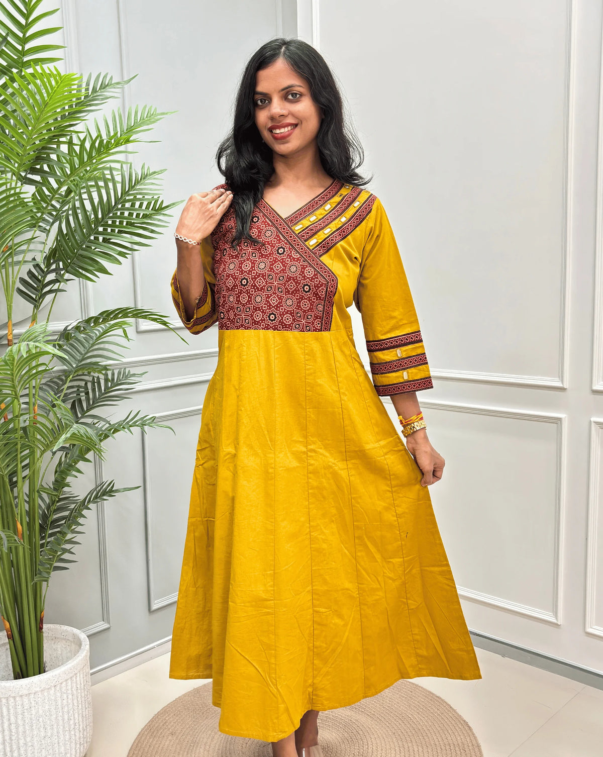 Front view of Yellow Women's Ajrakh Pure Cotton A-line Anarkali with a Red base & Black floral yoke patterned along with Real Hand-mirror work on Front & sleeve cuffs, standing on a round woven rug in front of a light gray paneled wall.