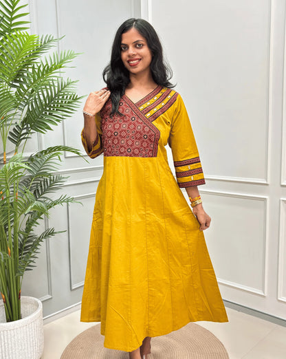 Front view of Yellow Women's Ajrakh Pure Cotton A-line Anarkali with a Red base & Black floral yoke patterned along with Real Hand-mirror work on Front & sleeve cuffs, standing on a round woven rug in front of a light gray paneled wall.