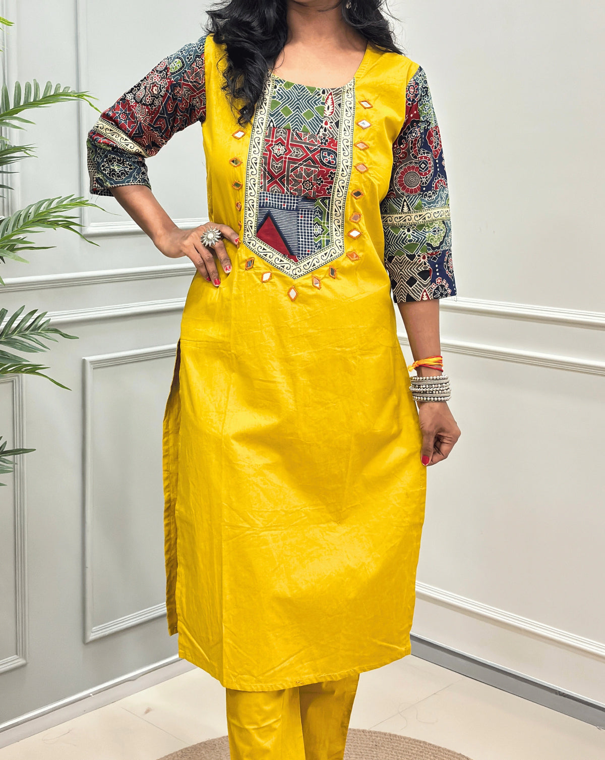Close-up of Yellow women's Ajrakh Vegetable dyed plant based Cotton Co-Ord set Featured with Round neck & Three-quarter sleeves with Multi-colour ajrakh Print detailed embroidered on neckline & Sleeves with beige strip border on work , Hand-mirror work out-line on front, paired with matching teal pants, standing on a round woven mat in a bright room with a potted plant.