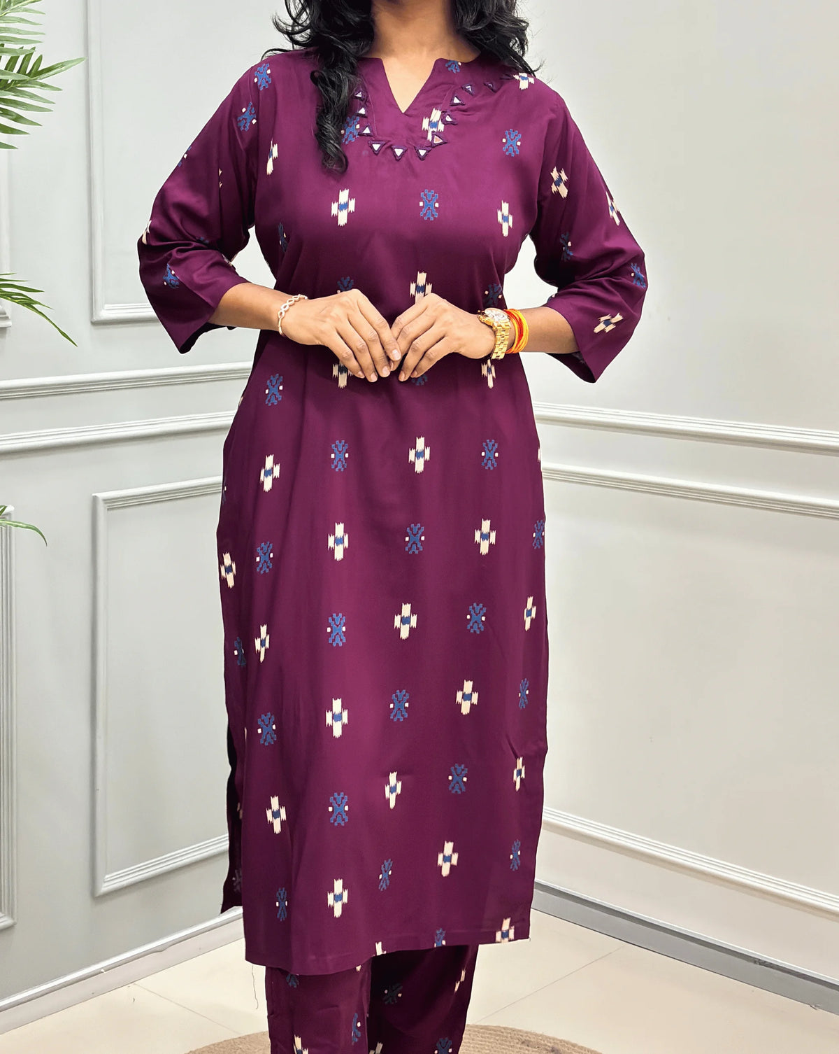 Close-up View of Purple Women's Ajrakh Vegetable Dyed Cotton Long Co-Ord Set Featured with Round V-neck & Three-Quarter Sleeves with white and blue geometric print & Real Hand-Mirror Work on Neck-line, along with Same Matching pant, standing on a round woven mat in front of a white paneled wall with a green plant on the left.