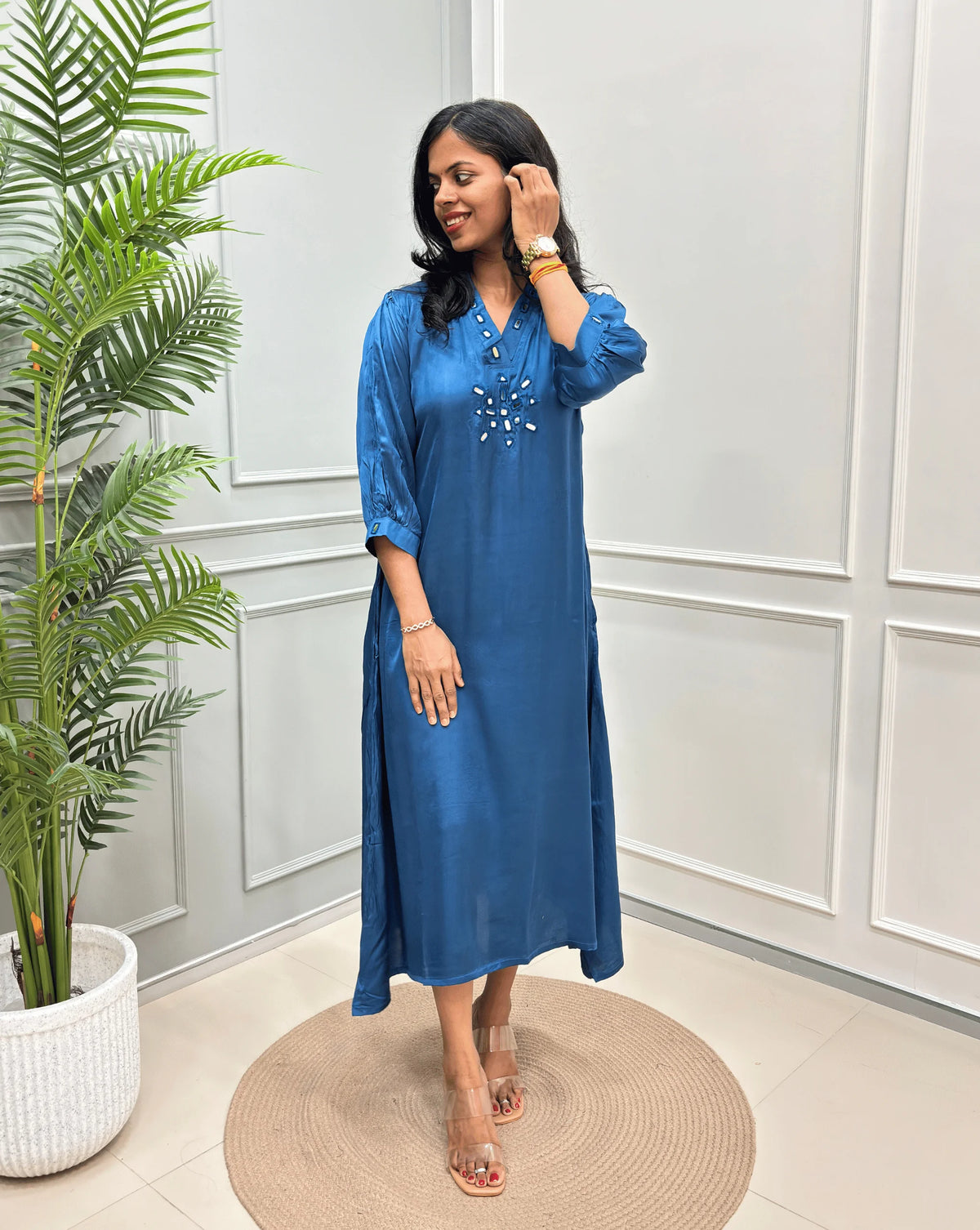 Blue Women's Pure Modal Silk Ajrakh Long Kurti with a decorative Real Hand-Mirror embroidered neckline, Floral pattern on front & Sleeves Cuffs, standing on a round woven rug in front of a white paneled wall with a potted plant on the left