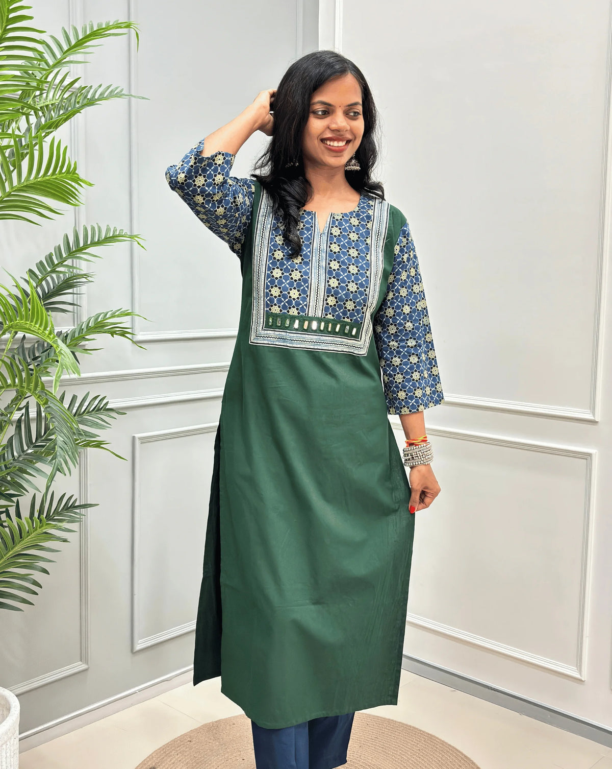 Front view of Dark Green Women's Hand-block Ajrakh Pure Cotton Long Kurti with a blue base , an abstract of black & beige Floral Print yoke on front & Full sleeves, Hand-Mirror work Front, paired with blue Pant, standing on a round woven mat in front of a white paneled wall.