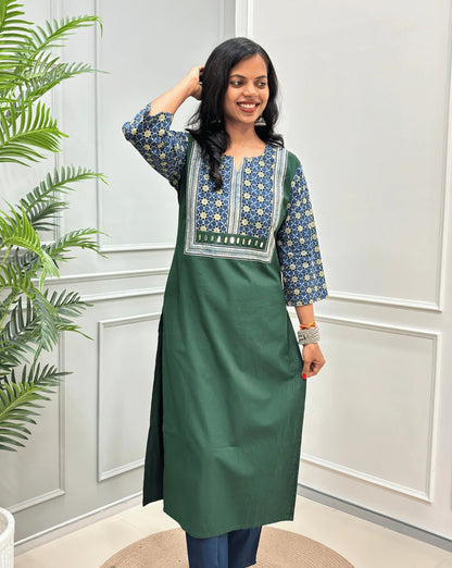 Front view of Dark Green Women's Hand-block Ajrakh Pure Cotton Long Kurti with a blue base , an abstract of black & beige Floral Print yoke on front & Full sleeves, Hand-Mirror work Front, paired with blue Pant, standing on a round woven mat in front of a white paneled wall.