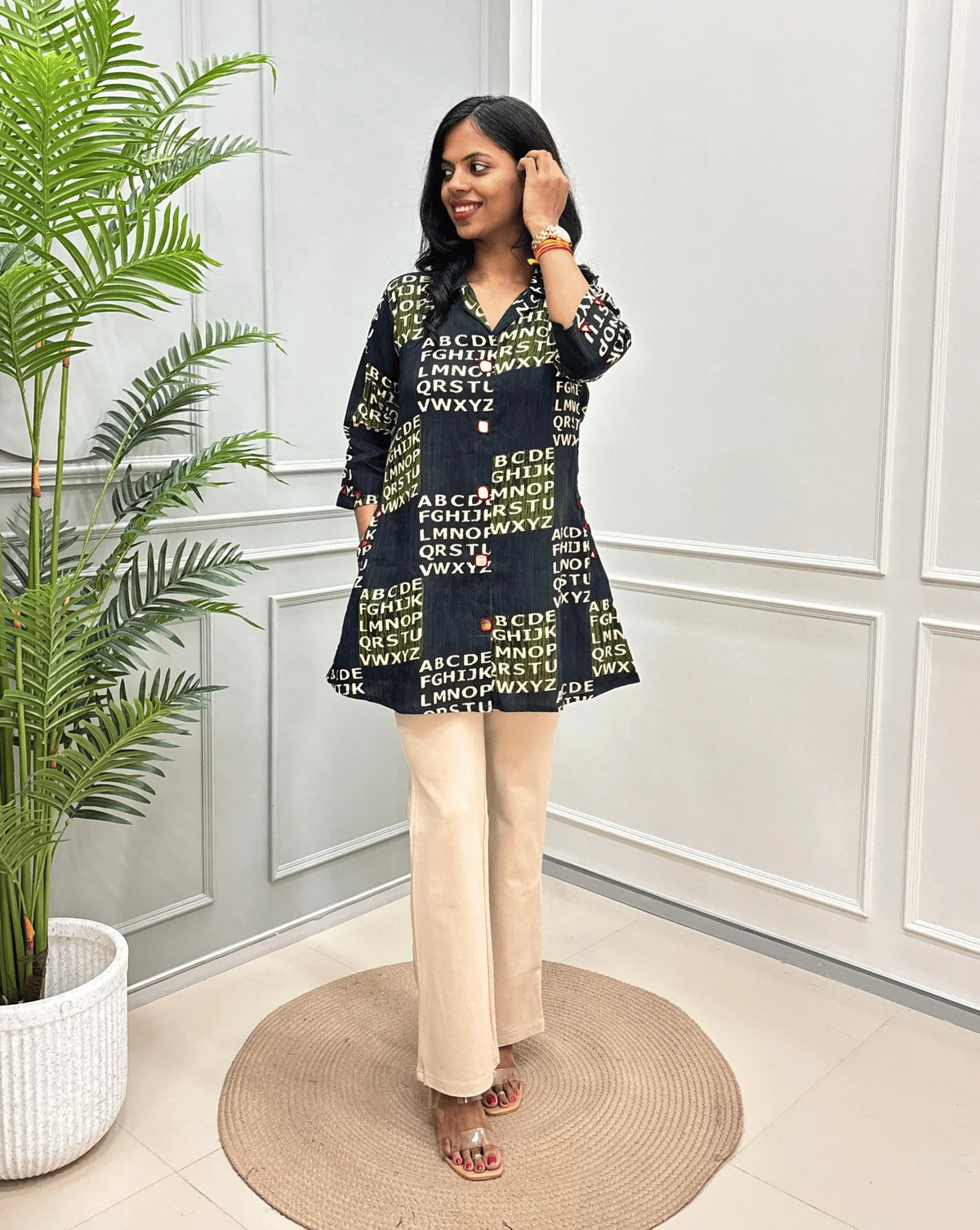 Green Women's Ajrakh Vegetable Dyed Plant based Cotton Short kurti with a Abstract Beige color alphabet print, paired with beige trousers, standing on a round woven rug in front of a white paneled wall with a potted plant.