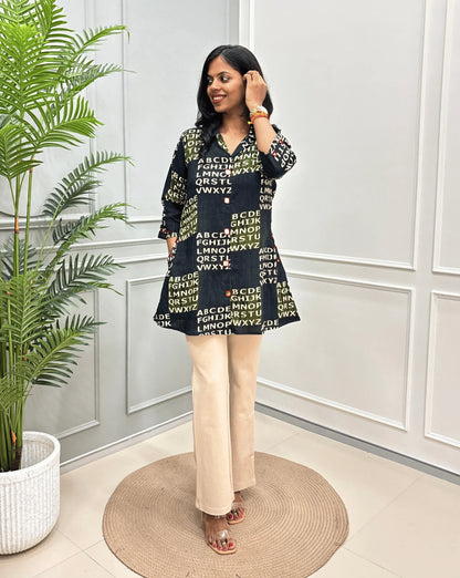 Green Women's Ajrakh Vegetable Dyed Plant based Cotton Short kurti with a Abstract Beige color alphabet print, paired with beige trousers, standing on a round woven rug in front of a white paneled wall with a potted plant.