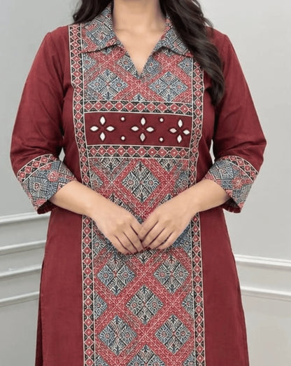 Close-up of Maroon Women's Ajrakh Vegetable Dyed Plant Based Cotton Long-Kurti Featured with Full pattren Collar V shaped Neck along with Three quarter Sleeves, with a Red Base, an Abstract of Black & Beige geometric patterned panel on front & Sleeves Cuffs, Real Hand-Mirror Patch work on Front, paired with Red pants, standing in a bright room with a white & Grey paneled wall, on left side a potted plant.