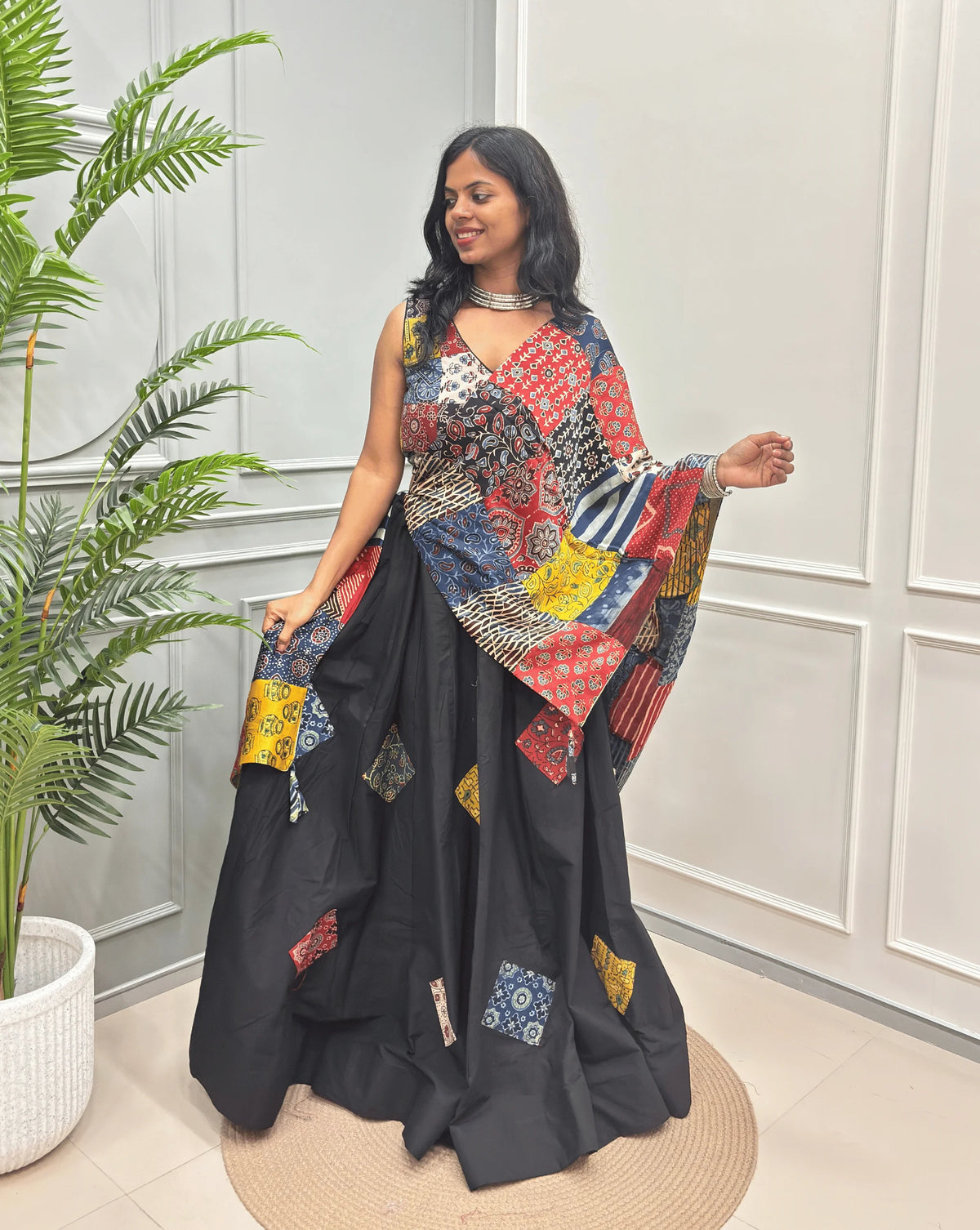 Black Women's Ajrakh pure Cotton Skirt Set with a beautiful Tasals & Ajrakh Patch work along with multi-colour Printed blouse & Dupatta, Standing against a light paneled wall with a green plant on the left.