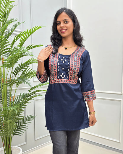 Close-up of Mashru-silk Short kurti detailed featuring with red, blue & red ajrakh print & mirror embroidery on neck-line, standing in front of a white paneled wall next to a green potted plant on left-side.