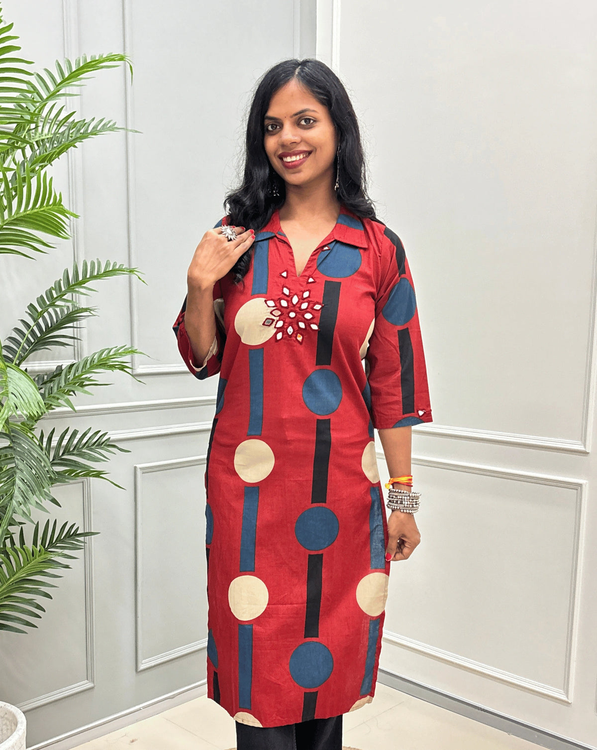 Front-view of Red Women's Ajrakh Vegetable dyed plant based Cotton Long-Kurti with a geometric pattern of Blue, beige, and black circles, Hand-mirror work on front in flower shape & Sleeves cuffs,  paired with black Flared pant, standing in a room with a potted plant and paneled walls.