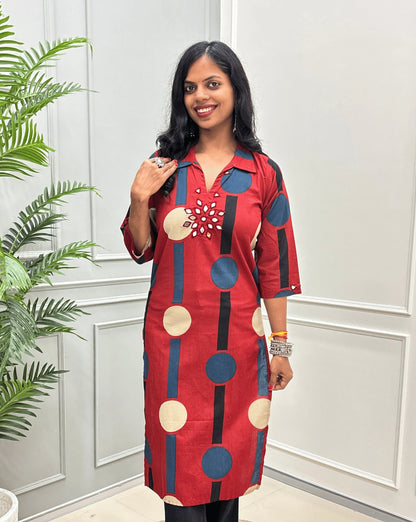 Front-view of Red Women's Ajrakh Vegetable dyed plant based Cotton Long-Kurti with a geometric pattern of Blue, beige, and black circles, Hand-mirror work on front in flower shape & Sleeves cuffs,  paired with black Flared pant, standing in a room with a potted plant and paneled walls.