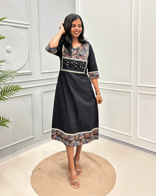 Black Women's Ajrakh Pure Cotton Long Kurti with a colorful Floral Printed yoke on  Chest, Cuffs  and hem, standing on a round woven rug against a white paneled wall.