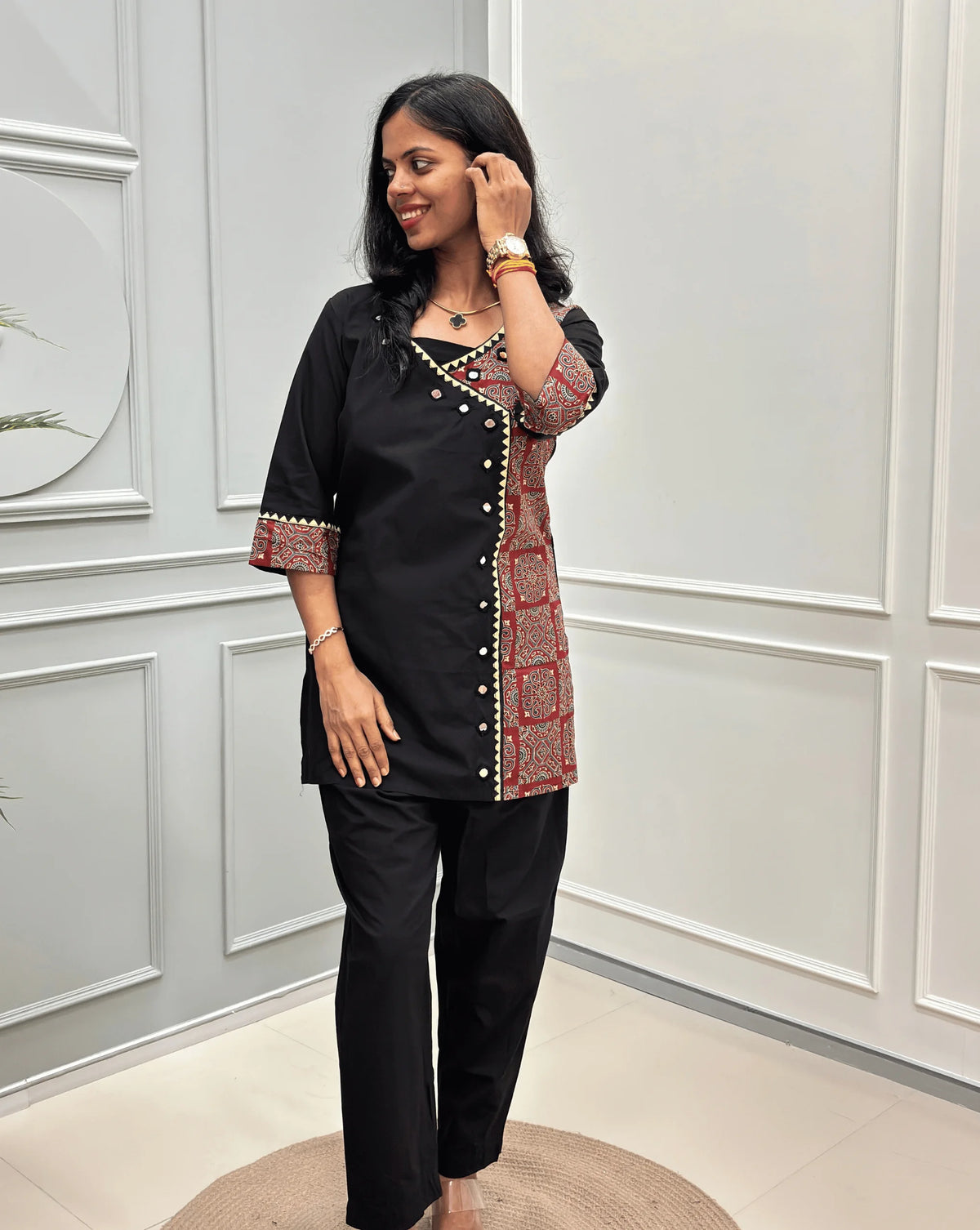 Front-view of Black Women's Ajrakh Pure Cotton 2-peice Co-Ord set with a Ajrakh printed panel on the front & Sleeves, Angrakha Patterned on front, Real Hand-Mirror work on front Along with 2 side pockets in pant, standing on a round woven rug in a modern room with white paneled walls.