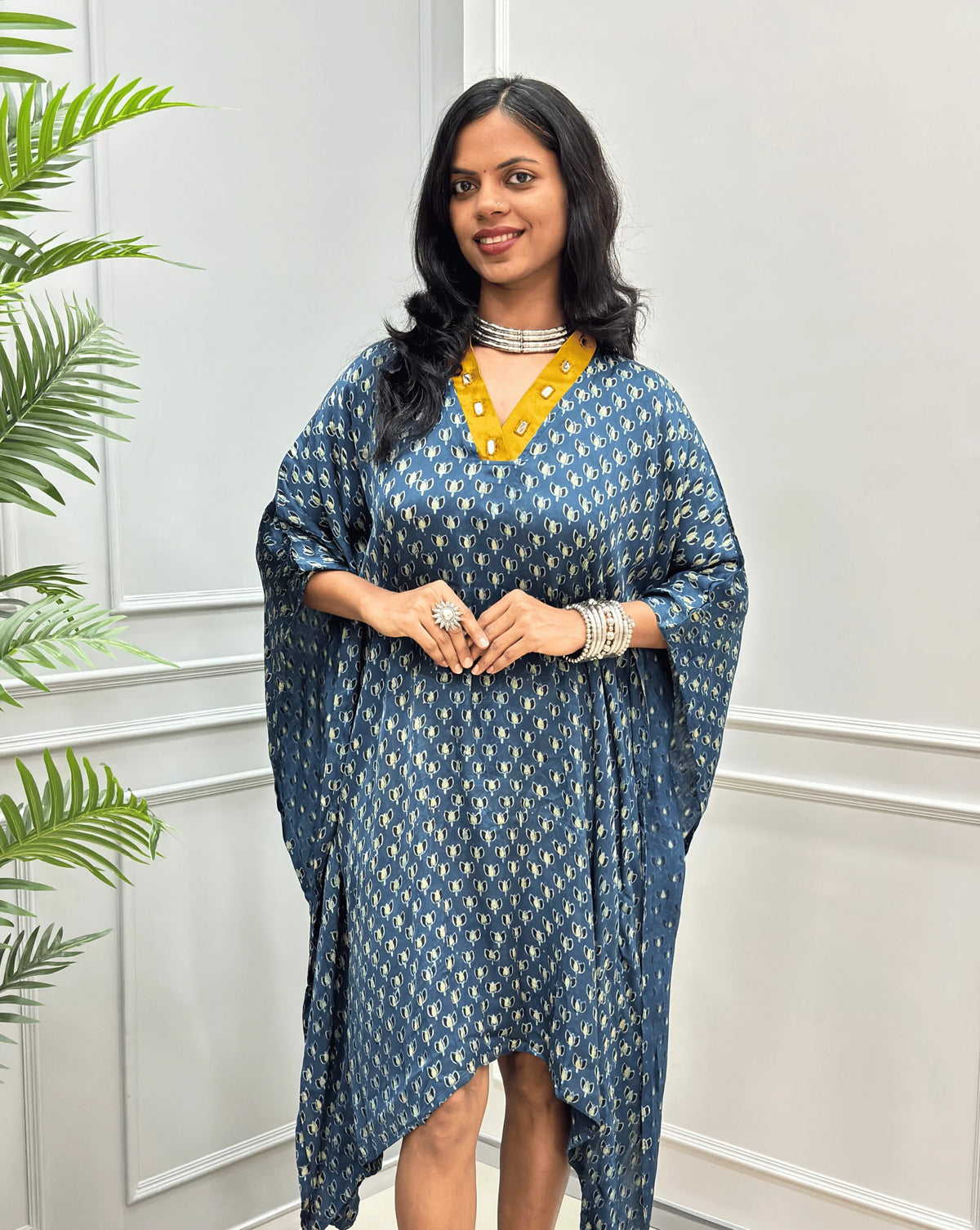 Front-view of Blue Women's Ajrakh Pure Modal Silk Knee Length Kaftan with a black Floral print, with Contrast yellow Colour V-neck, standing on a round woven rug in front of a white paneled wall next to a potted plant. 