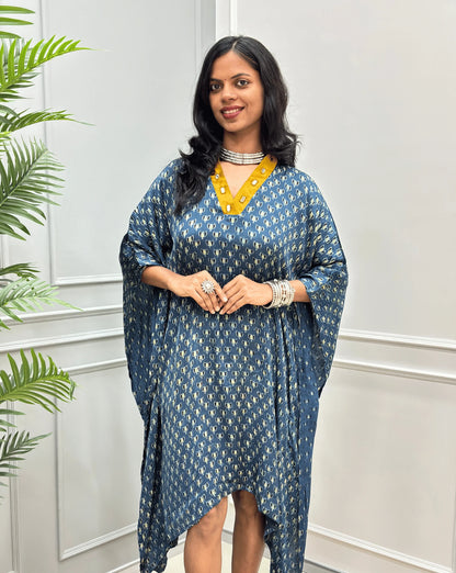 Front-view of Blue Women's Ajrakh Pure Modal Silk Knee Length Kaftan with a black Floral print, with Contrast yellow Colour V-neck, standing on a round woven rug in front of a white paneled wall next to a potted plant. 