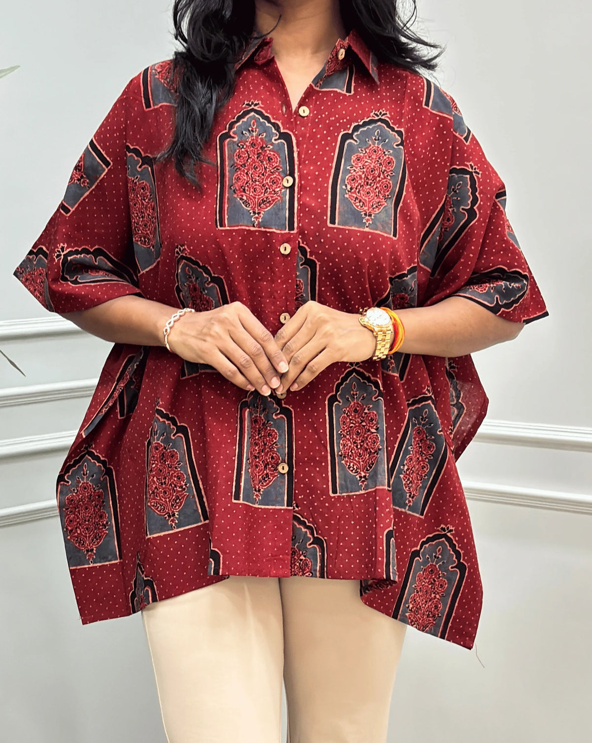 Close-up View of Red Women's Ajrakh Pure Cotton Short Kaftan Featured with Shirt Collar neck with a Black, Grey & Red Floral Block Print, Wooden Button on front, paired with beige flared pants, standing on a round woven rug in front of a white paneled wall with a potted plant.