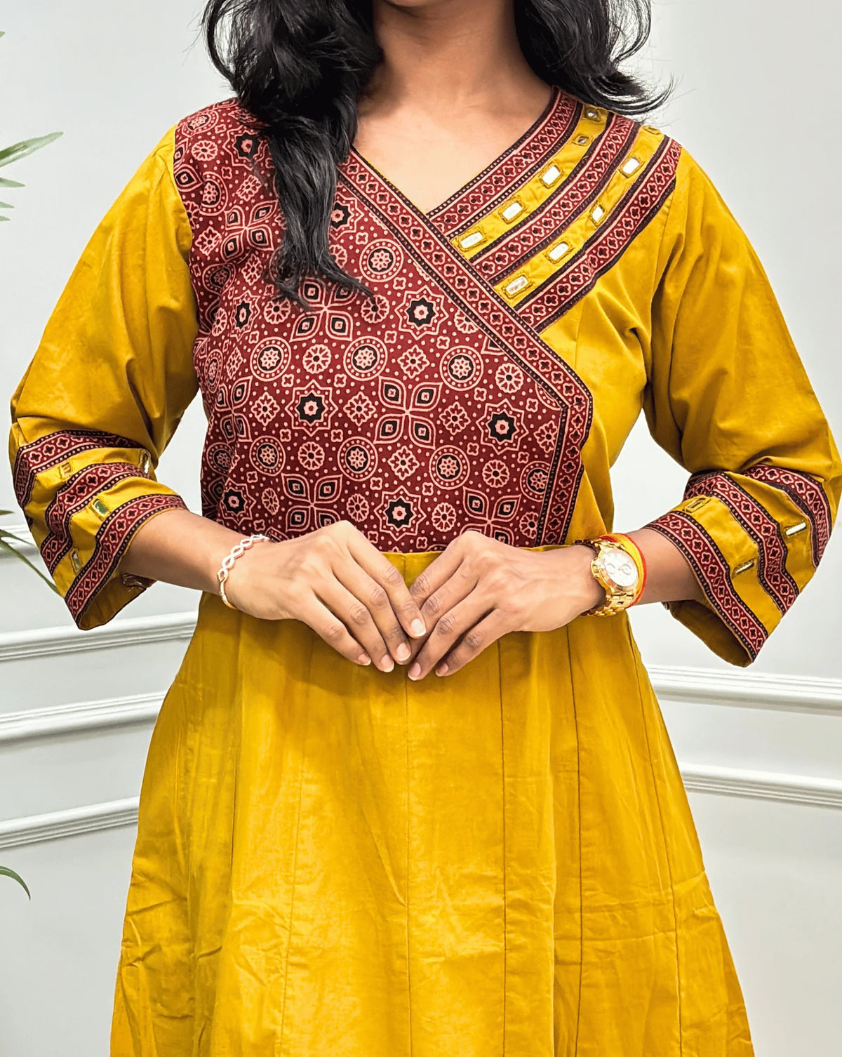 Close-up  view of Yellow Women's Ajrakh Pure Cotton A-line Anarkali Featured with  V-neck with a Red base & Black floral yoke patterned along with Real Hand-mirror work on Front & sleeve cuffs & 2-Side pockets, standing on a round woven rug in front of a light gray paneled wall.