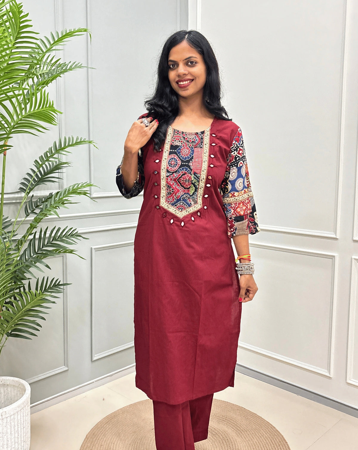 Front-view of Brick Red women's Ajrakh Vegetable dyed plant based Cotton Co-Ord set with Multi-colour ajrakh Print detailed embroidered on neckline & Sleeves with beige strip border on work , Hand-mirror work out-line on front, paired with matching teal pants, standing on a round woven mat in a bright room with a potted plant.