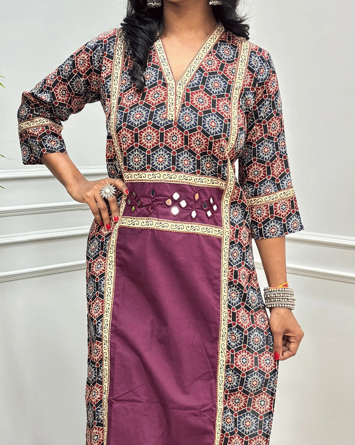 Close-up of Purple Women's Ajrakh pure cotton 2 peice Co-Ord set featured with V Shaped neck with a black base & an abstract of blue & Red Geometric print on Front Chest to Side & Sleeves , Hand-mirror work on Front as Belt, standing on a round woven mat in front of a white paneled wall with a potted plant.