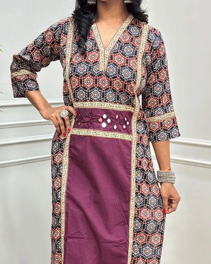 Close-up of Purple Women's Ajrakh pure cotton 2 peice Co-Ord set featured with V Shaped neck with a black base & an abstract of blue & Red Geometric print on Front Chest to Side & Sleeves , Hand-mirror work on Front as Belt, standing on a round woven mat in front of a white paneled wall with a potted plant.