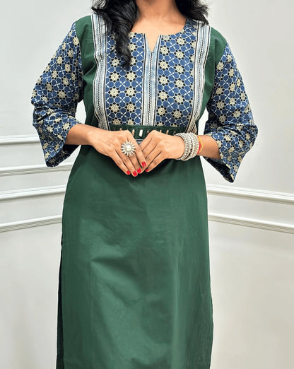 Close-up view of Dark Green Women's Hand-block Ajrakh Pure Cotton Long Kurti featured with Round V Shaped neck along with three quarter sleeves, with a blue base , an abstract of black & beige Floral Print yoke on front & Full sleeves, Hand-Mirror work Front, paired with blue Pant, standing on a round woven mat in front of a white paneled wall.