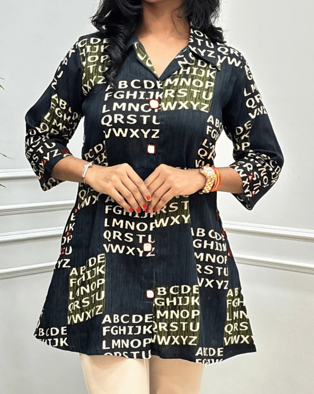 Close-up of Green Women's Ajrakh Vegetable Dyed Plant based Cotton Short kurti featured with Collar neck & Three Quarter Sleeves, with a Abstract Beige color alphabet print, Hand-mirror work on Front, Sleeves & pockets, paired with beige trousers, standing on a round woven rug in front of a white paneled wall with a potted plant.