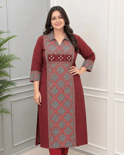Front-Side View of Maroon Women's Ajrakh Vegetable Dyed Plant Based Cotton Long-Kurti with a Red Base,  an Abstract of Black, blue & Beige geometric patterned panel on front & Sleeves Cuffs, Real Hand-Mirror Patch work on Front, paired with Red pants, standing in a bright room with a white & Grey paneled wall, on left side a potted plant.