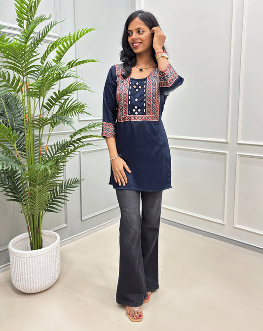 Navy Blue women's mashru silk short kurti with red and white embroidered details, paired with dark gray flare jeans and clear flat sandals, standing in a room with a potted plant.