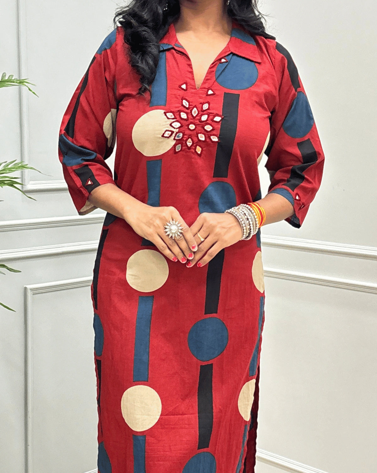 Close-up of Dark Red Women's Ajrakh Vegetable dyed plant based Cotton Long-Kurti Featured Collar V-neck & Three-quarter Sleeves with a geometric pattern of Blue, beige, and black circles, Hand-mirror work on front in flower shape & Sleeves cuffs, paired with black Flared pant, standing in a room with a potted plant and paneled walls.
