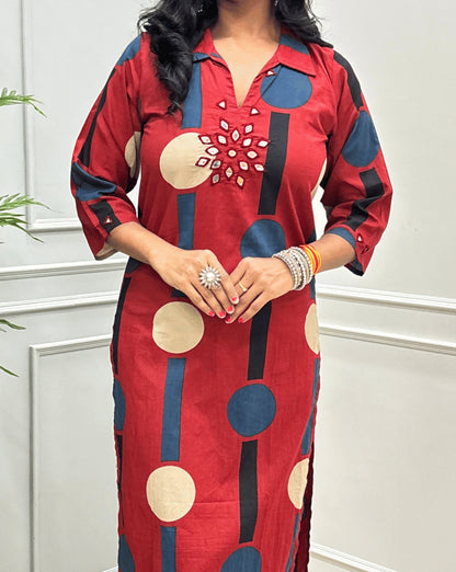 Close-up of Dark Red Women's Ajrakh Vegetable dyed plant based Cotton Long-Kurti Featured Collar V-neck & Three-quarter Sleeves with a geometric pattern of Blue, beige, and black circles, Hand-mirror work on front in flower shape & Sleeves cuffs, paired with black Flared pant, standing in a room with a potted plant and paneled walls.