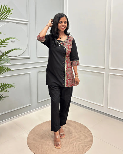 Black Women's Ajrakh Pure Cotton 2-peice Co-Ord set with a Ajrakh printed panel on the front & Sleeves, Angrakha Patterned on front, standing on a round woven rug in a modern room with white paneled walls.