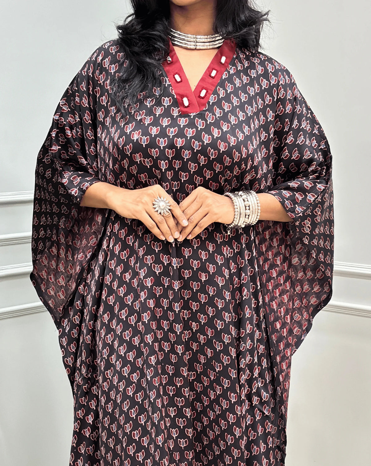 Close-up view of Black Women's Ajrakh Pure Modal Silk Knee Length Kaftan with a black Floral print, with Contrast Red Colour V-neck, along with Real Hand-Mirror embroidery, standing on a round woven rug in front of a white paneled wall next to a potted plant. 