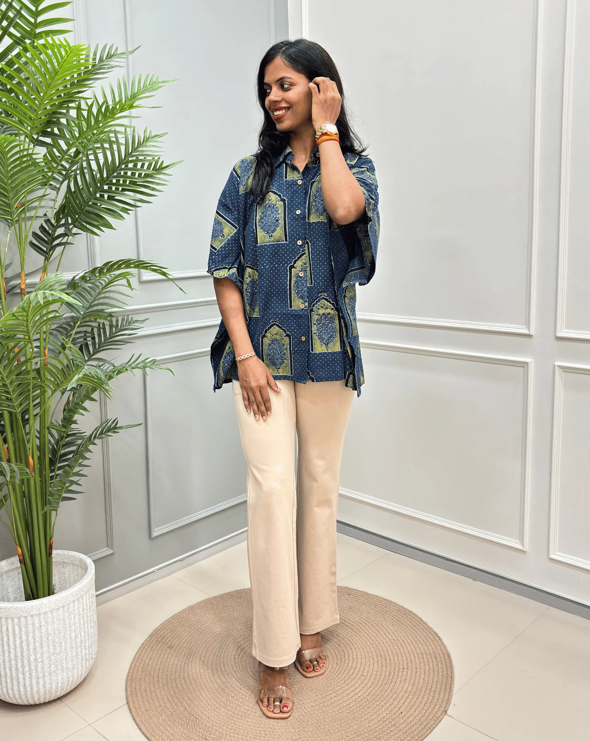 Blue Women's Ajrakh Pure Cotton Short Kaftan Shirt with a Black, Green & Yellow Floral Block Print, paired with beige flared pants, standing on a round woven rug in front of a white paneled wall with a potted plant.