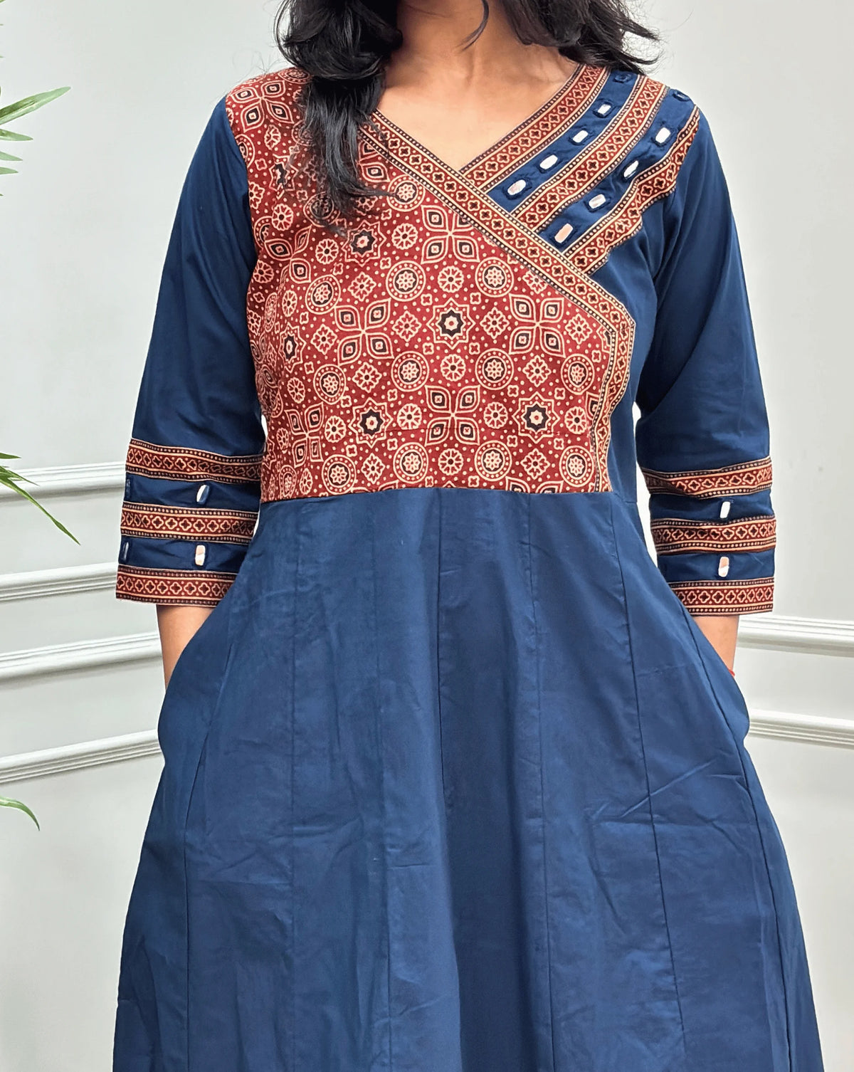 Close-up view of Navy-blue Women's Ajrakh Pure Cotton A-line Anarkali featured with V-neck with a Red base & Black floral yoke patterned along with Real hand-mirror work on Front & sleeve cuffs & 2-side pockets , standing on a round woven rug in front of a light gray paneled wall.