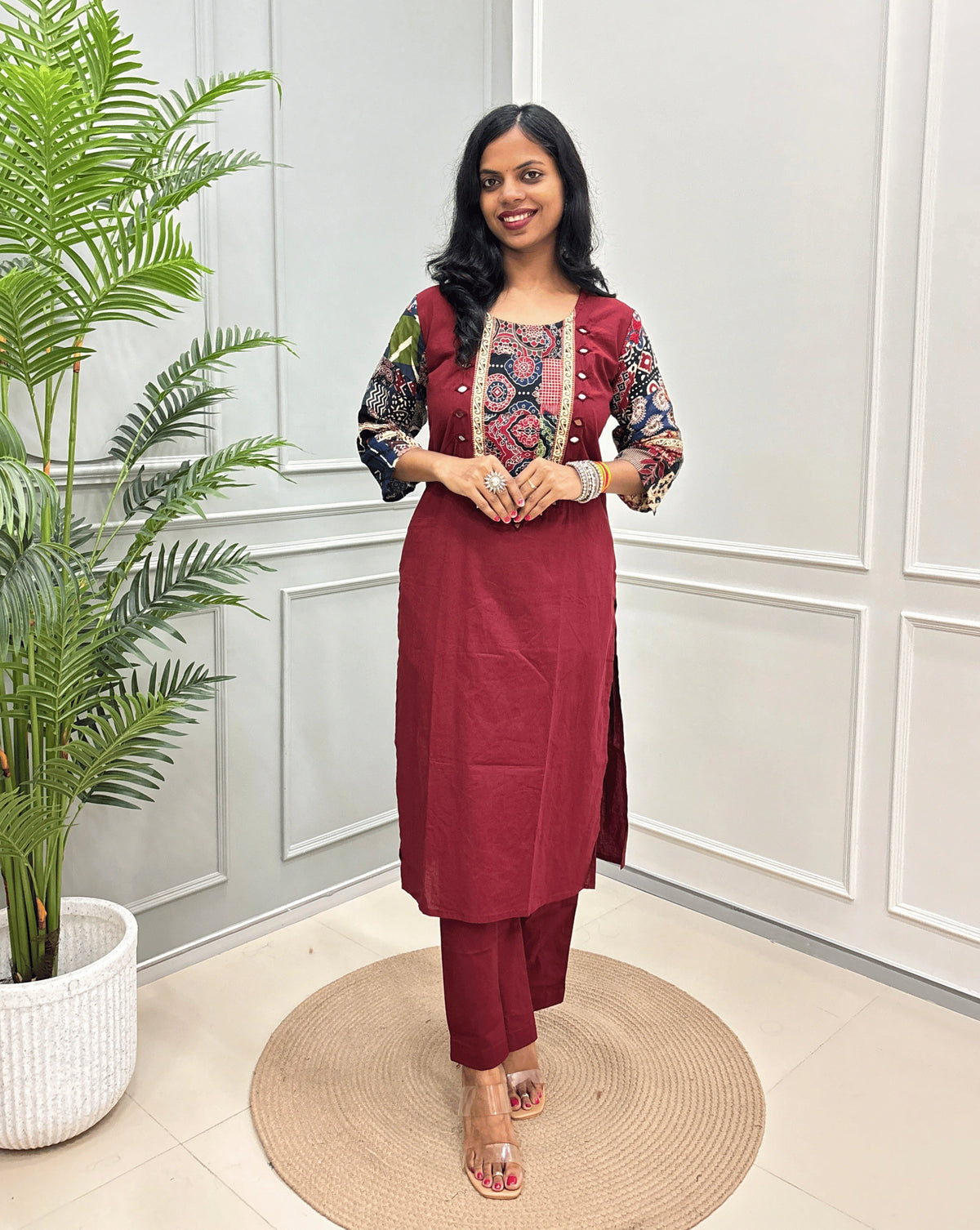 Brick Red women's Ajrakh Vegetable dyed plant based Cotton CoOrd set with Multi-colour ajrakh Print detailed embroidered on neckline & Sleevess with beige border on work ,  paired with matching teal pants, standing on a round woven mat in a bright room with a potted plant.