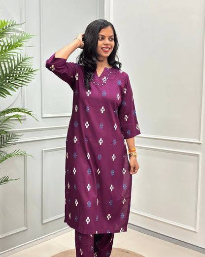 Front-view of Purple Women's Ajrakh Vegetable Dyed Cotton Long Co-Ord Set with white and blue geometric print & Real Hand-Mirror Work on Neck-line, along with Same Matching pant, standing on a round woven mat in front of a white paneled wall with a green plant on the left.