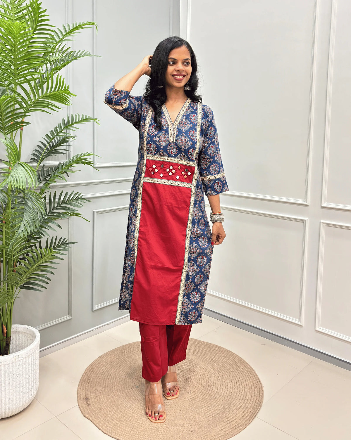 Red Women's Ajrakh pure cotton 2 peice Co-Ord set with a Blue base & an Peacock print on Front Chest to Side & Sleeves , standing on a round woven mat in front of a white paneled wall with a potted plant.
