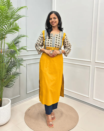 Mustard Yellow Women's Hand-block Ajrakh Pure Cotton Long Kurti with a Beige base , an abstract of Blue & Black Floral Print yoke on front & Full sleeves, paired with blue Pant, standing on a round woven mat in front of a white paneled wall.