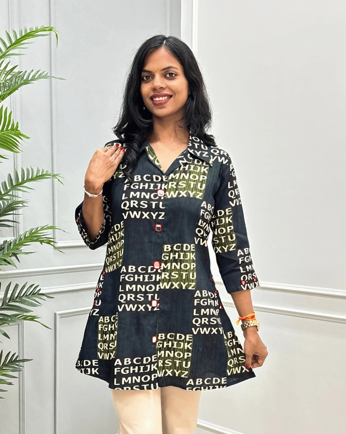 Front View of Green Women's Ajrakh Vegetable Dyed Plant based Cotton Short kurti with a Abstract Beige color alphabet print, Hand-mirror work on Front, Sleeves & pockets, paired with beige trousers, standing on a round woven rug in front of a white paneled wall with a potted plant.