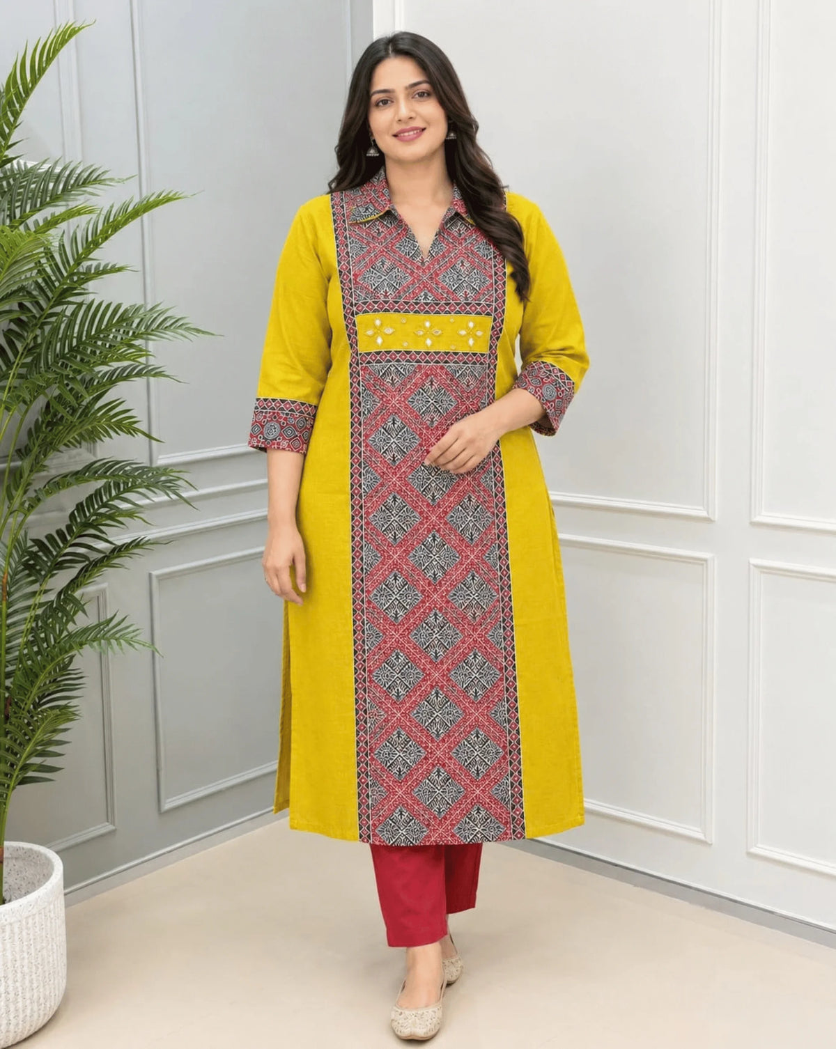 Mustard Yellow Women's Ajrakh Vegetable Dyed Plant Based Cotton Long-Kurti with a red Base,  an Abstract of Black  & Beige geometric patterned panel on front & Sleeves Cuffs, paired with red pants, standing in a bright room with a white & Grey paneled wall ,on left side a potted plant.