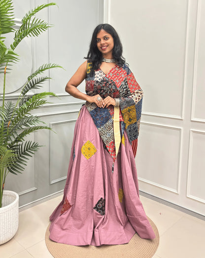 Pink Women's Ajrakh pure Cotton Skirt Set with a beautiful Tasals & Ajrakh Patch work along with multi-colour Printed blouse & Dupatta, Standing against a light paneled wall with a green plant on the left.