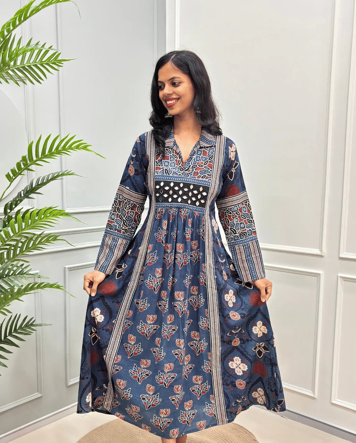 Blue cotton Ajrakh Long dress front view with intricate red, beige, and white floral and geometric patterns, Along with 2 side pockets, standing in front of a white paneled wall with a green plant on the left.