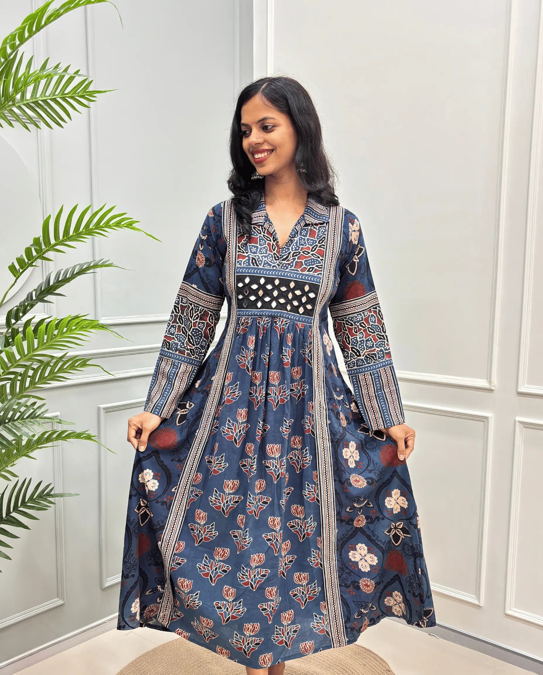Blue cotton Ajrakh Long dress front view with intricate red, beige, and white floral and geometric patterns, Along with 2 side pockets, standing in front of a white paneled wall with a green plant on the left.