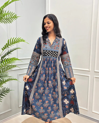Blue cotton Ajrakh Long dress front view with intricate red, beige, and white floral and geometric patterns, Along with 2 side pockets, standing in front of a white paneled wall with a green plant on the left.
