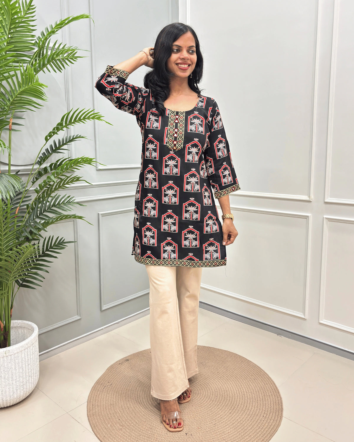 Black Women's Ajrakh vegetable Dyed Plant based Cotton Short Kurti with a colorful Pichhwai pattern, Paired with beige flared pants, standing on a round woven mat in front of a white paneled wall with a potted plant.