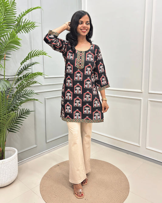 Black Women's Ajrakh vegetable Dyed Plant based Cotton Short Kurti with a colorful Pichhwai pattern, Paired with beige flared pants, standing on a round woven mat in front of a white paneled wall with a potted plant.