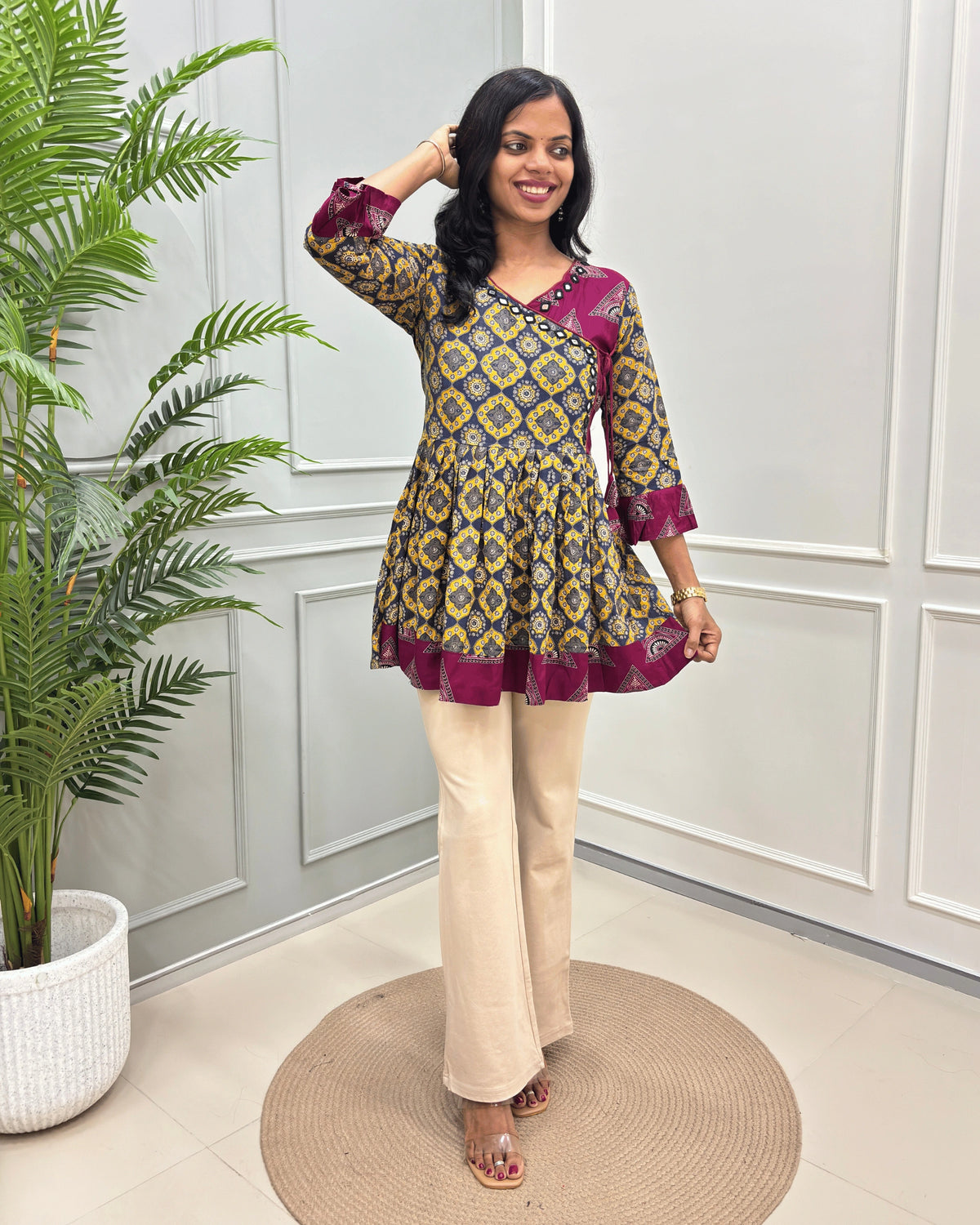 Yellow Women's Ajrakh Vegetable dyed Plant based Rayon Cotton with a yellow, blue, and deep pink geometric pattern, paired with beige wide-leg trousers, standing on a round woven mat in front of a white paneled wall.