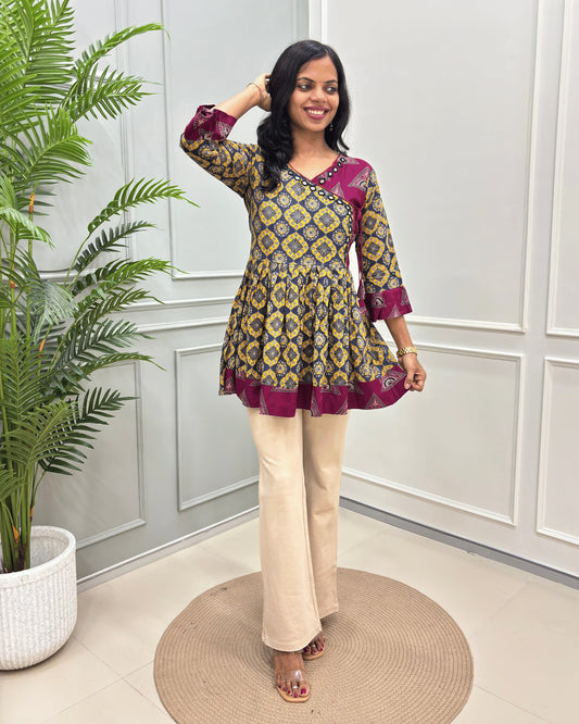 Yellow Women's Ajrakh Vegetable dyed Plant based Rayon Cotton with a yellow, blue, and deep pink geometric pattern, paired with beige wide-leg trousers, standing on a round woven mat in front of a white paneled wall.