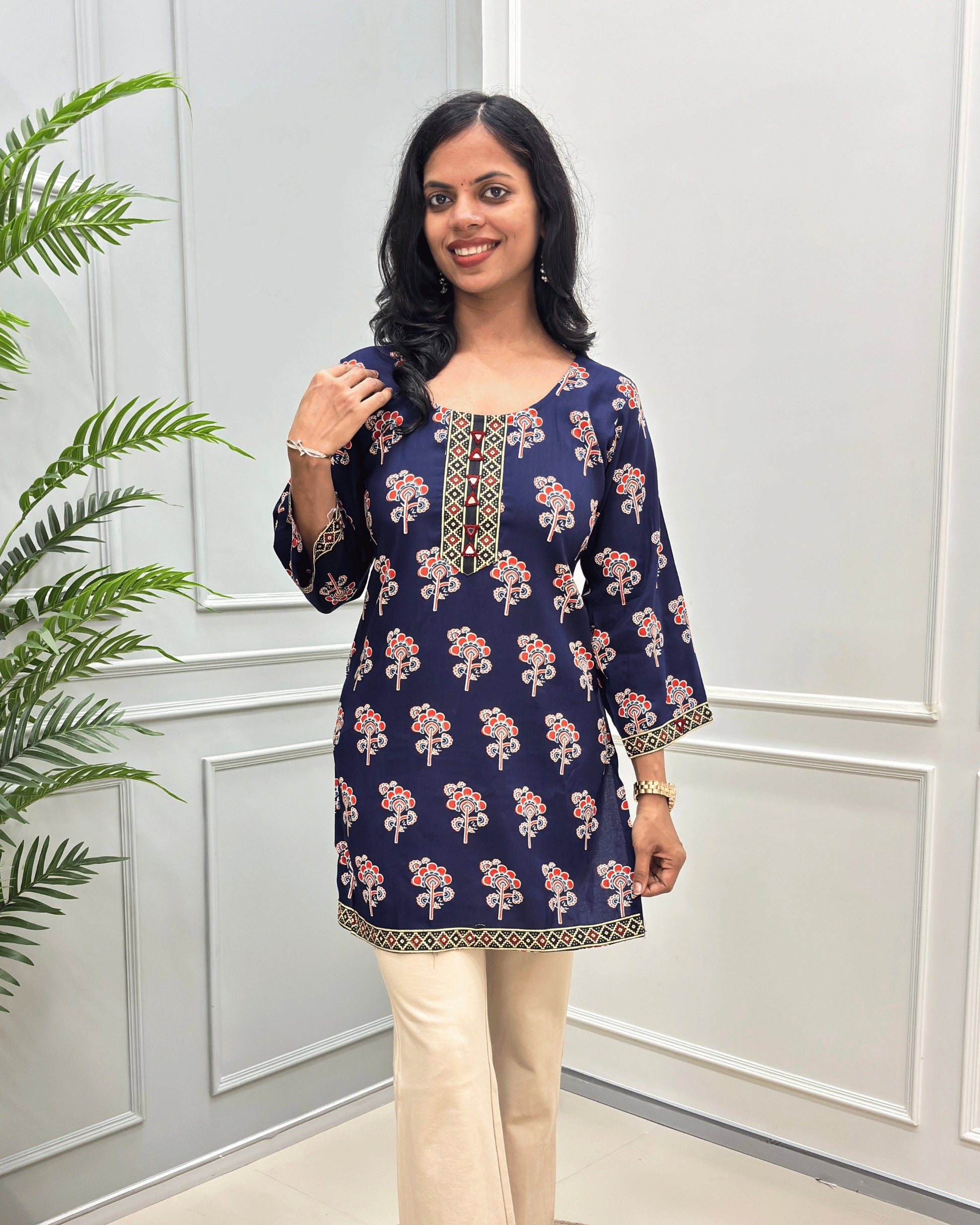 Front view of Navy-Blue women's Ajrakh plant based cotton Short kurti with a colorful floral print, paired with beige pant , standing in front of a white paneled wall with a green plant on the left. 