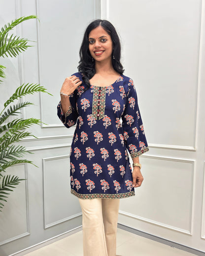 Front view of Navy-Blue women's Ajrakh plant based cotton Short kurti with a colorful floral print, paired with beige pant , standing in front of a white paneled wall with a green plant on the left. 
